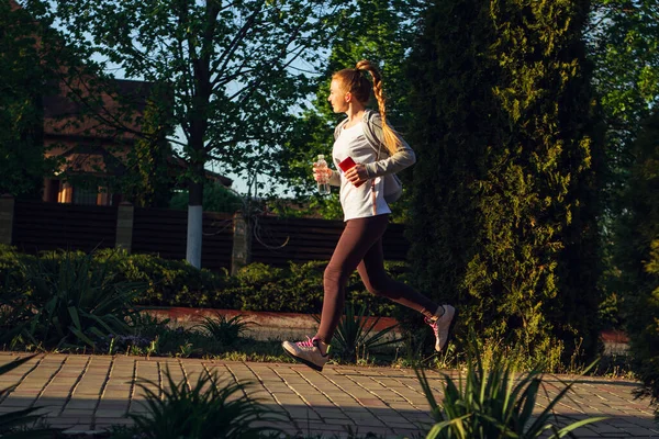 Joven corredora, atleta está corriendo en la calle de la ciudad bajo el sol. Hermosa mujer caucásica entrenando, escuchando música —  Fotos de Stock