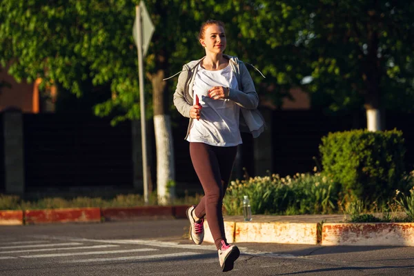 Jonge vrouwelijke loper, atleet is joggen in de stad straat in de zon. Mooie Kaukasische vrouw training, luisteren naar muziek — Stockfoto