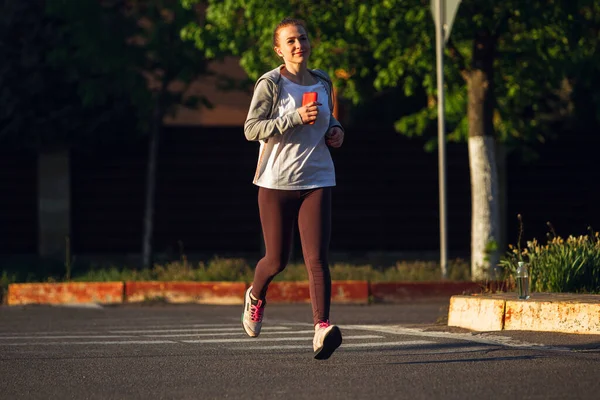 Joven corredora, atleta está corriendo en la calle de la ciudad bajo el sol. Hermosa mujer caucásica entrenando, escuchando música — Foto de Stock