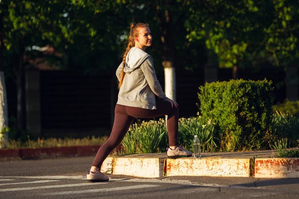 Joven corredora, atleta se está estirando antes de correr en la calle de la ciudad bajo el sol. Hermosa mujer caucásica entrenando, escuchando música — Foto de Stock