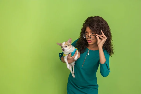 Afro-Amerikalı güzel genç kadın portresi yeşil stüdyo arka planında küçük köpekli, duygusal ve anlamlı. Reklam için kopyalama alanı. — Stok fotoğraf