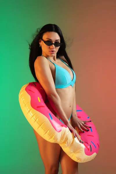 Modeporträt einer jungen, fitten und sportlichen Frau in blauer Luxus-Badebekleidung mit Gummi-Donut und stylischer Sonnenbrille auf Steigungsuntergrund. Perfekter Körper bereit für den Sommer. — Stockfoto