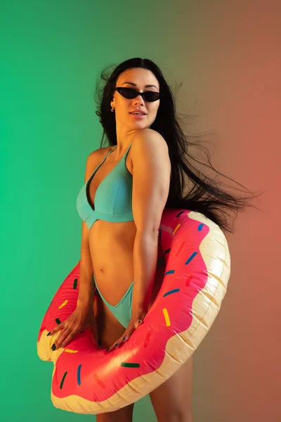 Retrato de moda de mulher jovem em forma e esportiva em roupa de banho de luxo azul com donut de borracha e óculos de sol elegantes no fundo gradiente. Corpo perfeito pronto para o verão . — Fotografia de Stock