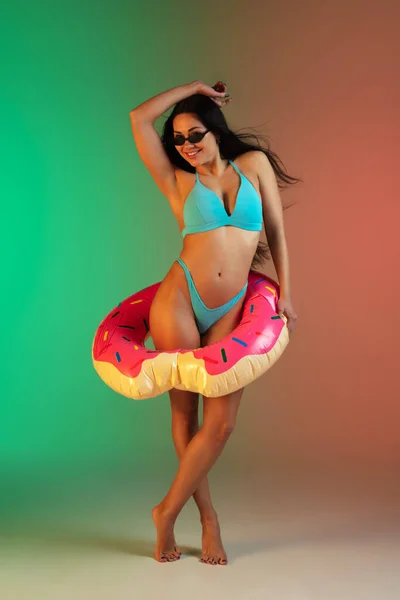 Retrato de moda de mulher jovem em forma e esportiva em roupa de banho de luxo azul com donut de borracha e óculos de sol elegantes no fundo gradiente. Corpo perfeito pronto para o verão . — Fotografia de Stock