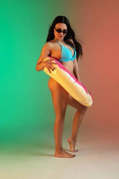 Modeporträt einer jungen, fitten und sportlichen Frau in blauer Luxus-Badebekleidung mit Gummi-Donut und stylischer Sonnenbrille auf Steigungsuntergrund. Perfekter Körper bereit für den Sommer. — Stockfoto