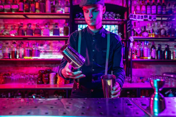 Barman finishes preparation of alcoholic cocktail with shaker in multicolored neon light