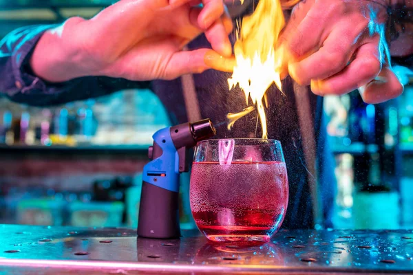 Primer plano de cóctel alcohólico, bebida, bebida en luz de neón multicolor con llamas de fuego encendidas en el jugo de pulverización de naranjas — Foto de Stock