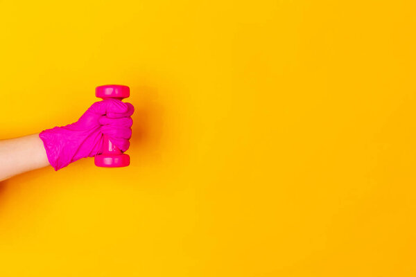 Hand in pink rubber glove holding dumbbell, gym weight isolated on yellow studio background with copyspace.