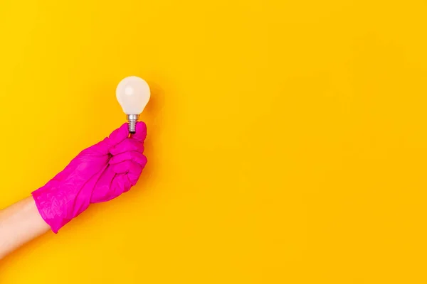 Main en gant de caoutchouc rose tenant ampoule isolée sur fond de studio jaune avec copyspace . — Photo