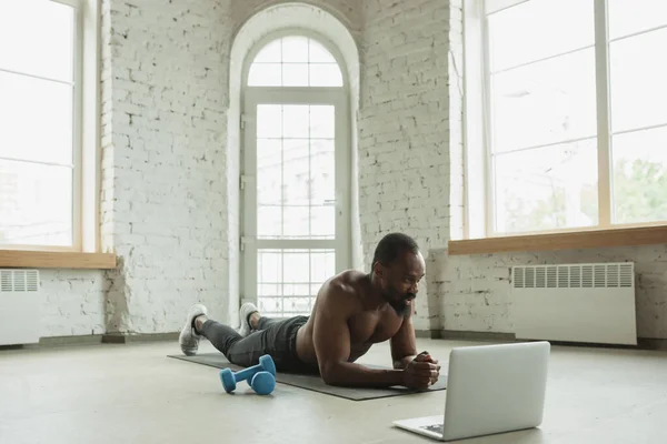 Hombre afroamericano joven entrenando en casa durante la cuarentena del brote de coronavirus, ejercicios de doinc de fitness, aeróbicos. Mantener el aislamiento deportivo asegurando . — Foto de Stock