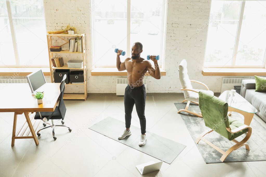 Young african-american man training at home during quarantine of coronavirus outbreak, doinc exercises of fitness, aerobic. Staying sportive suring insulation.