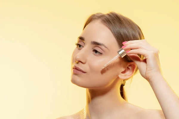 Bescherming. Close-up van mooie vrouwelijke gezicht met gieten etherische olie, serum over gele achtergrond. Cosmetica en make-up, natuurlijke en eco-behandeling, huidverzorging. — Stockfoto