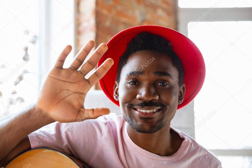 African-american musician in red hat greeting audience before online concert at home isolated and quarantined, cheerful, smiling