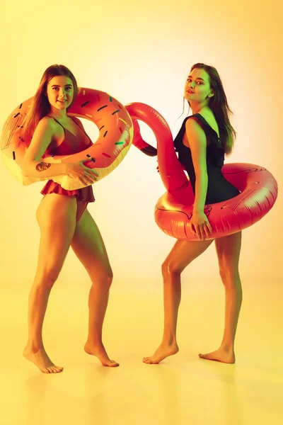 Schöne Mädchen in modischen Badeanzügen isoliert auf gelbem Studiohintergrund in Neonlicht. Sommer, Resort, Mode und Wochenendkonzept. Posieren in Gummi-Donut und Flamingo mit hellen Emotionen. — Stockfoto