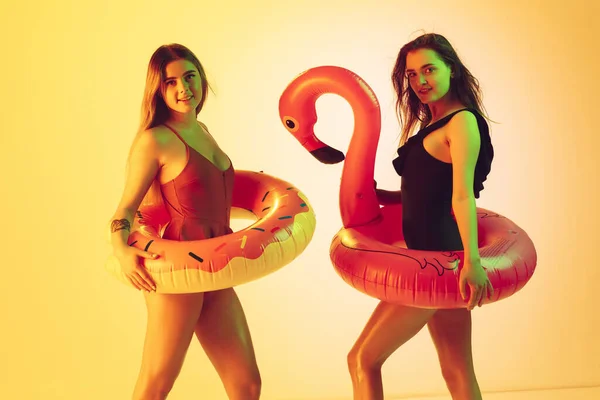 Schöne Mädchen in modischen Badeanzügen isoliert auf gelbem Studiohintergrund in Neonlicht. Sommer, Resort, Mode und Wochenendkonzept. Posieren in Gummi-Donut und Flamingo mit hellen Emotionen. — Stockfoto