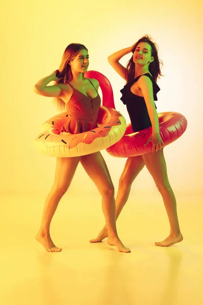 Hermosas chicas en trajes de baño de moda aislados en el fondo del estudio amarillo en luz de neón. Verano, resort, moda y concepto de fin de semana. Posando en donut de goma y flamenco con emociones brillantes . — Foto de Stock