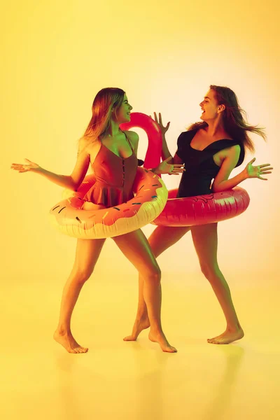 Hermosas chicas en trajes de baño de moda aislados en el fondo del estudio amarillo en luz de neón. Verano, resort, moda y concepto de fin de semana. Posando en donut de goma y flamenco con emociones brillantes . —  Fotos de Stock