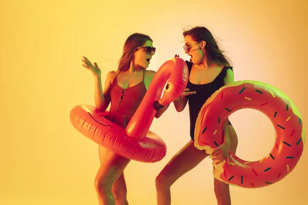 Hermosas chicas en trajes de baño de moda aislados en el fondo del estudio amarillo en luz de neón. Verano, resort, moda y concepto de fin de semana. Posando en donut de goma y flamenco con emociones brillantes . — Foto de Stock