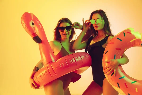 Meninas bonitas em trajes de banho elegantes isolados no fundo do estúdio amarelo em luz de néon. Verão, resort, moda e conceito de fim de semana. Posando em donut de borracha e flamingo com emoções brilhantes . — Fotografia de Stock