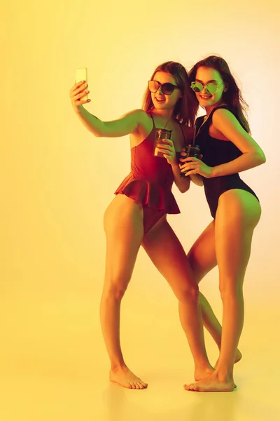 Meninas bonitas em trajes de banho elegantes isolados no fundo do estúdio amarelo em luz de néon. Verão, resort, moda e conceito de fim de semana. Tomar selfie com coquetéis . — Fotografia de Stock