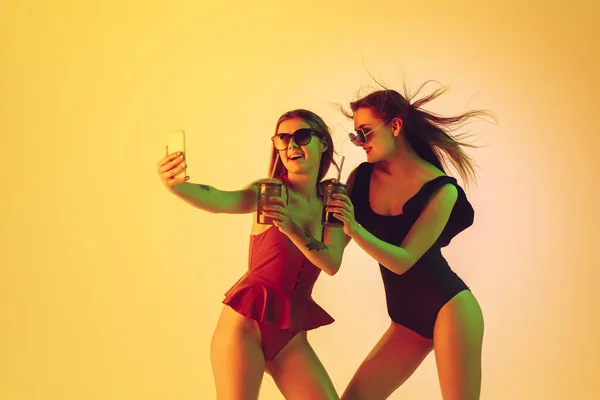 Hermosas chicas en trajes de baño de moda aislados en el fondo del estudio amarillo en luz de neón. Verano, resort, moda y concepto de fin de semana. Tomar selfie con cócteles . —  Fotos de Stock