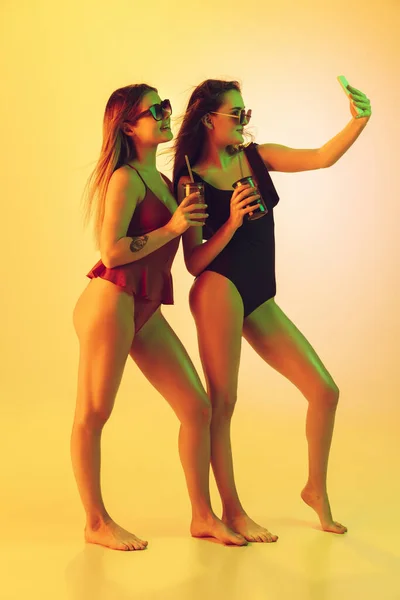 Hermosas chicas en trajes de baño de moda aislados en el fondo del estudio amarillo en luz de neón. Verano, resort, moda y concepto de fin de semana. Tomar selfie con cócteles . — Foto de Stock