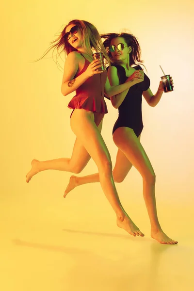 Красиві дівчата в модних купальниках ізольовані на жовтому студійному фоні в неоновому світлі. Літо, курорт, мода та концепція вихідних. З коктейлями на бігу . — стокове фото