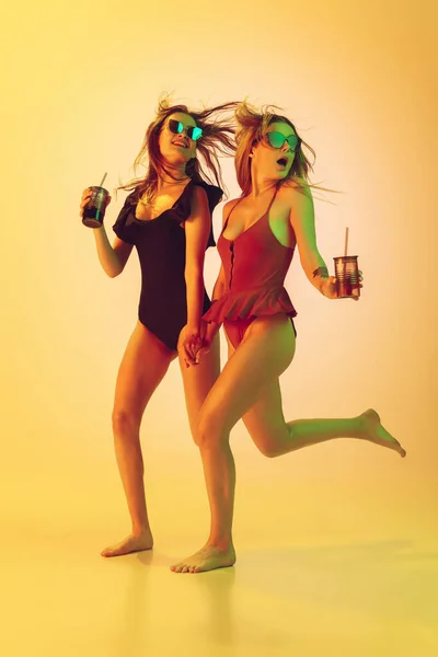 Schöne Mädchen in modischen Badeanzügen isoliert auf gelbem Studiohintergrund in Neonlicht. Sommer, Resort, Mode und Wochenendkonzept. Mit Cocktails auf der Flucht. — Stockfoto