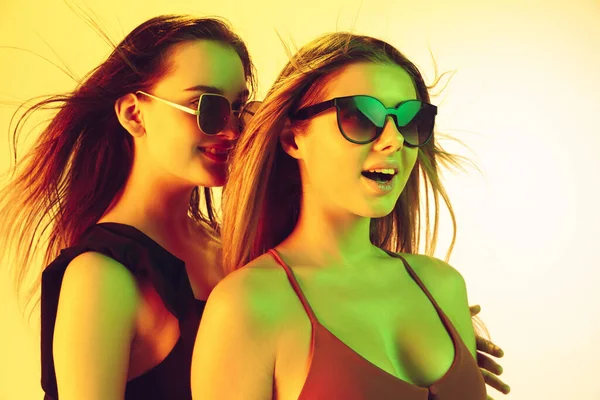 Close up beautiful girls in fashionable swimsuits and eyewear isolated on yellow studio background in neon light. Summer, resort, fashion and weekend concept. Stock Picture