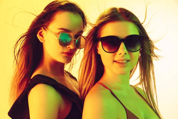 Close up beautiful girls in fashionable swimsuits and eyewear isolated on yellow studio background in neon light. Summer, resort, fashion and weekend concept. Stock Photo