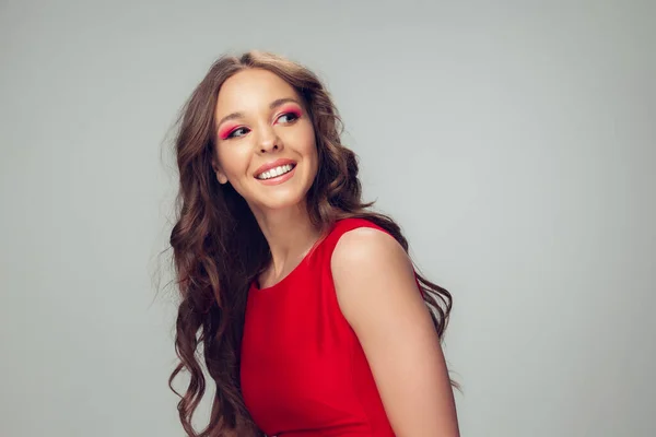 Belle jeune femme aux longs cheveux bouclés sains et au maquillage lumineux portant une robe rouge isolée sur fond de studio gris. Souriant . — Photo