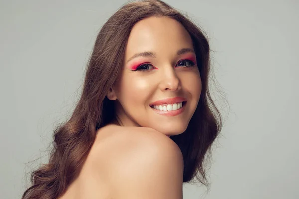 Primer plano de la hermosa mujer joven con el pelo rizado largo y saludable y brillante maquillaje aislado en el fondo del estudio gris, tímido sonriendo —  Fotos de Stock