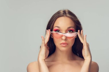 Uzun, sağlıklı kıvırcık saçlı, parlak makyajlı, gri stüdyo arka planında izole edilmiş pembe gözlüklü güzel bir genç kadın.