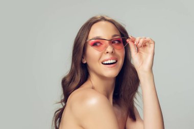 Uzun, sağlıklı kıvırcık saçlı, parlak makyajlı, şık pembe gözlüklü, gri stüdyo arka planında izole edilmiş, gülümseyen güzel bir genç kadın.