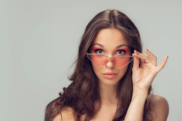 Κοντινό πλάνο της όμορφης νεαρής γυναίκας με μακριά υγιή σγουρά μαλλιά και φωτεινό μακιγιάζ φορώντας κομψό ροζ γυαλιά που απομονώνονται σε γκρι στούντιο backgroud — Φωτογραφία Αρχείου