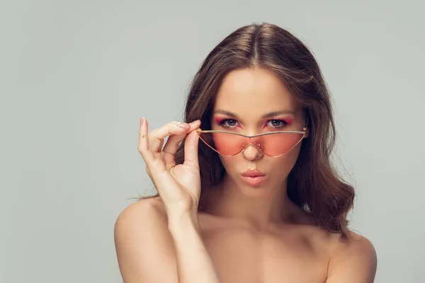 Close-up van mooie jonge vrouw met lang gezond krullend haar en heldere make-up dragen stijlvolle roze brillen geïsoleerd op grijze studio backgroud, blazen — Stockfoto