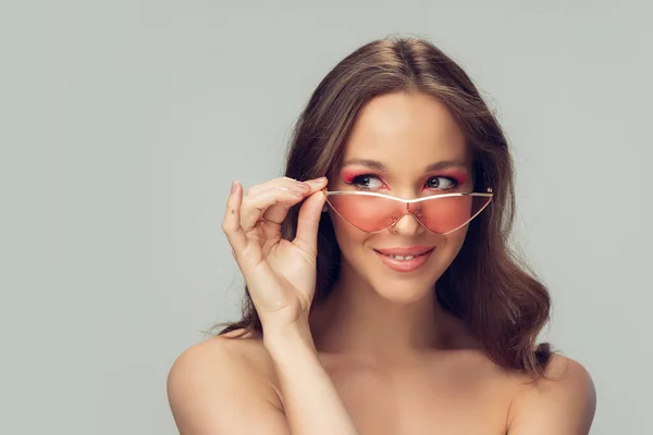 Primer plano de la hermosa mujer joven con el pelo rizado largo y saludable y maquillaje brillante usando gafas de color rosa con estilo aislado en el fondo del estudio gris, sonriendo —  Fotos de Stock