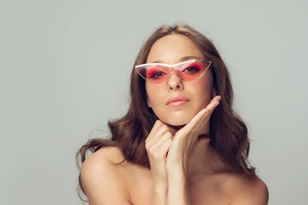 Close-up van mooie jonge vrouw met lang gezond krullend haar en heldere make-up dragen stijlvolle roze brillen geïsoleerd op grijze studio backgroud — Stockfoto
