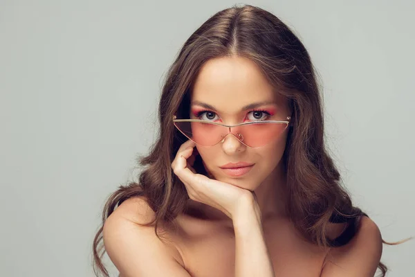 Gros plan de belle jeune femme avec de longs cheveux bouclés en bonne santé et un maquillage lumineux portant des lunettes roses élégantes isolées sur fond studio gris — Photo