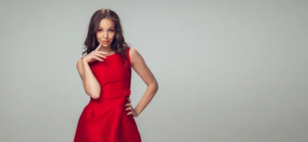 Hermosa mujer joven con pelo rizado largo y saludable y maquillaje brillante con vestido rojo aislado en el fondo del estudio gris. Folleto con copyspace . — Foto de Stock