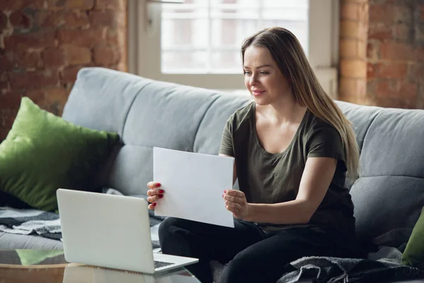 Young woman, businesswoman working or studying at home, looking on computer screen, monitor, holding white sheet, whiteboard. Attented, concentrated. Copyspace. Top view. — Stock Photo, Image