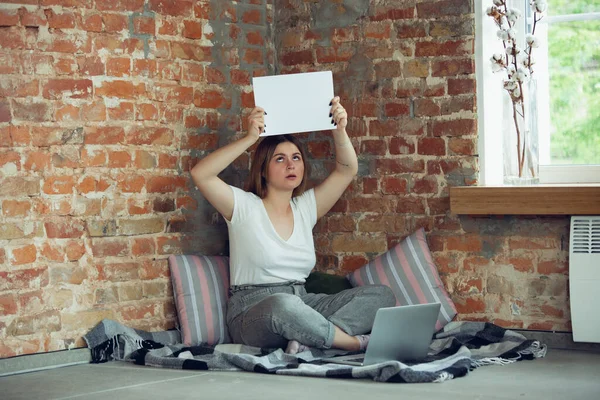 Молодая женщина, деловая женщина работает или учится дома, смотрит на экран компьютера, монитор, держит белый лист, доску. Акцентирован, сконцентрирован. Авторское пространство. Вид сверху . — стоковое фото