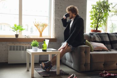 Pantolonsuz ama ceketli genç bir adam bilgisayar ve dizüstü bilgisayarla çalışıyor. Coronavirus sırasında uzak ofis, iç çamaşırlarıyla eğlenceli ve rahat bir iş.