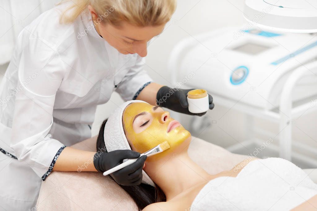Beautician applying radiant gold mask on face of beautiful woman.