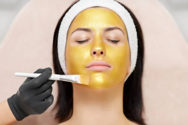 Cosmetologist applying gold mask on face of beautiful brunette. — Stock Photo, Image