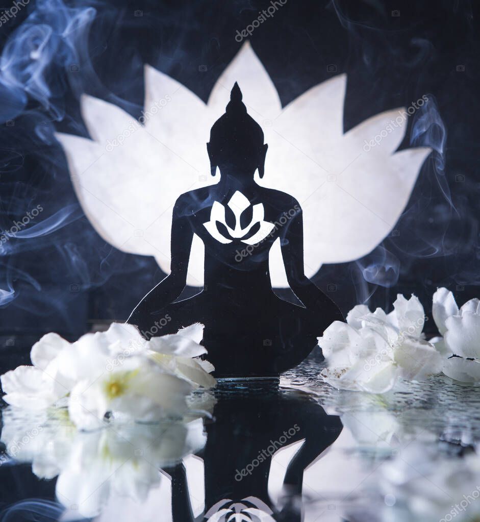 The photo shows the silhouette of a Buddha with a luminous lotus on his chest. The figure is surrounded by flowers. In the foreground, we see water and a reflection of the Buddha figure. On the background is a large lotus flower. 