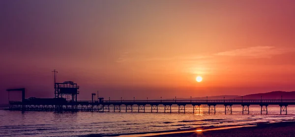 Photo of the sunset. A pier runs along the horizon. Photo taken in soft warm colors.