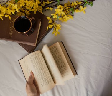 Fotoğrafta, bir kadın eli bir kitabın sayfalarını çeviriyor. Yakınlarda birkaç kitap, bir fincan kahve ve sarı çiçekler var. Kitabın sayfası biraz bulanık, bu da hareketlerini gösteriyor..