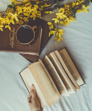 Fotoğrafta, bir kadın eli bir kitabın sayfalarını çeviriyor. Yakınlarda birkaç kitap, bir fincan koyu kahve ve sarı çiçekler var..