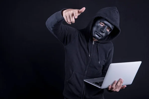 Ein anonymer männlicher Hacker mit schwarzer Maske bedeckt sein Gesicht, trägt einen Laptop und zeigt auf seine Hand. Konzepte für Internetsicherheit und Cyber-Angriffe. — Stockbild Hacker mit schwarzer Maske trägt Laptop — Stockfoto